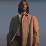 Keanu Reeves em pose contemplativa no filme "Quando o Céu se Engana", vestindo sobretudo claro sob o céu azul