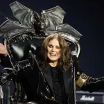 Ozzy Osbourne sentado em um trono com design de criatura alada, vestindo traje preto brilhante e braços abertos em pose teatral durante apresentação.