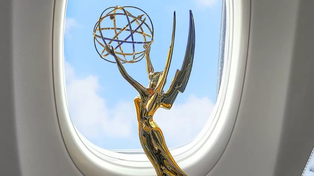 Uma estatueta dourada do Emmy, representando uma figura alada segurando um átomo, está posicionada próxima à janela de um avião. Ao fundo, vê-se o céu azul com algumas nuvens leves.