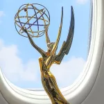 Uma estatueta dourada do Emmy, representando uma figura alada segurando um átomo, está posicionada próxima à janela de um avião. Ao fundo, vê-se o céu azul com algumas nuvens leves.